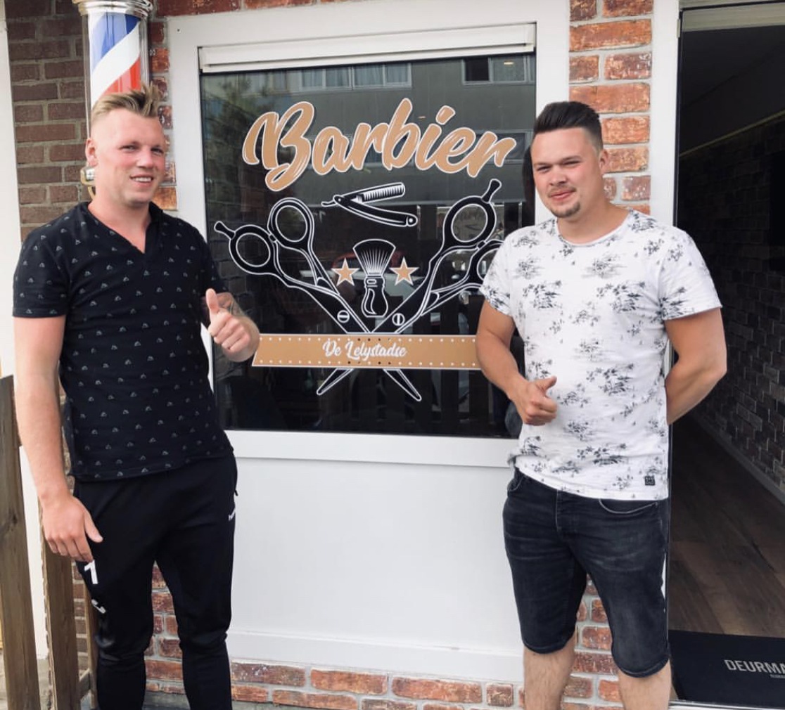 Twee mannen met de duim omhoog voor het raam van de zaak met de Barbier-belettering