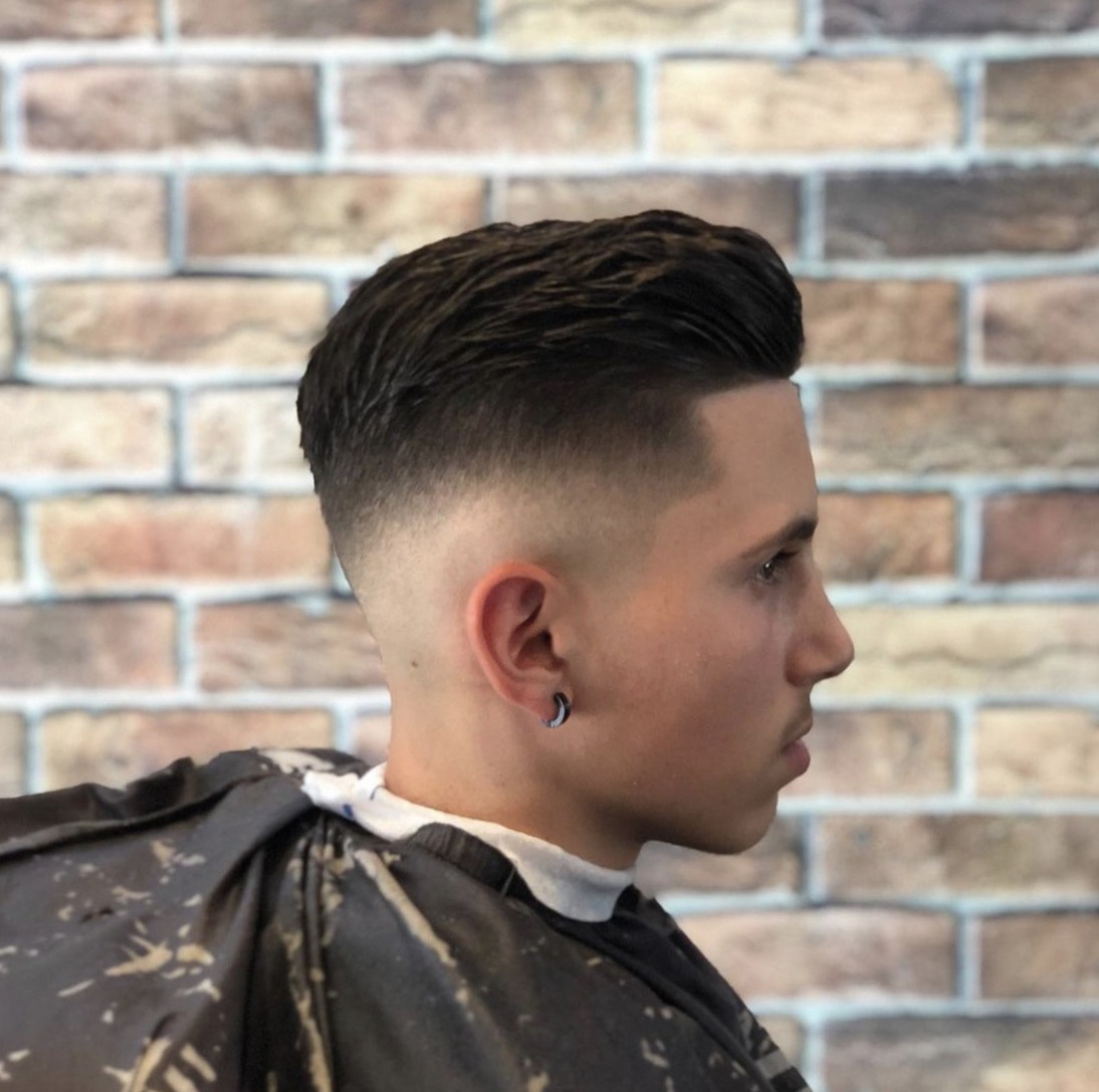 Skin fade met kuif, van opzij gezien tegen de bakstenen muur
