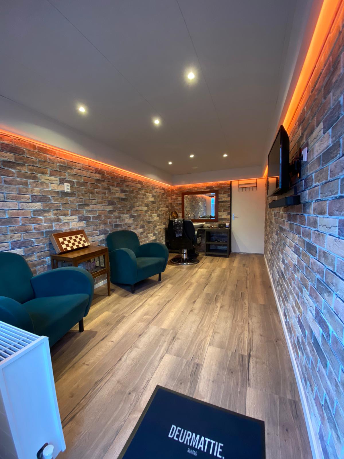 De zaak van binnen: bakstenen muur, ledverlichting, groene fauteuils en een houten vloer
